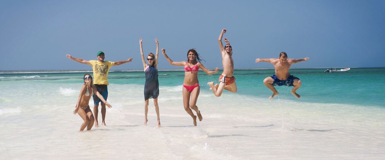 A group of friends jumping for joy on the white-sand shore of La Tortuga, with turquoise water behind